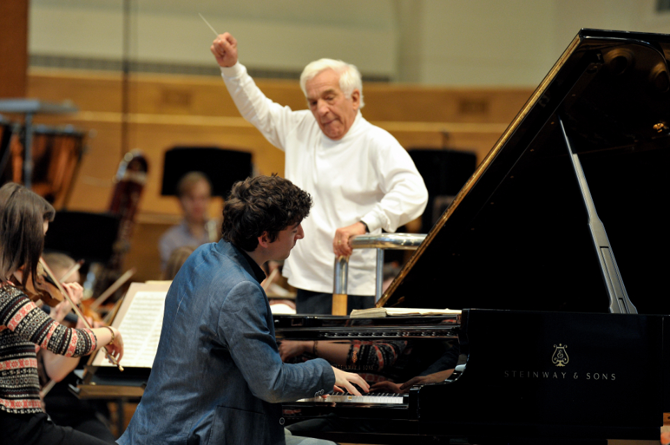 A male conductor, wearing a white shirt, with white hair, conducting an orchestra through a rehearsal, with a male pianist performing in the foreground.